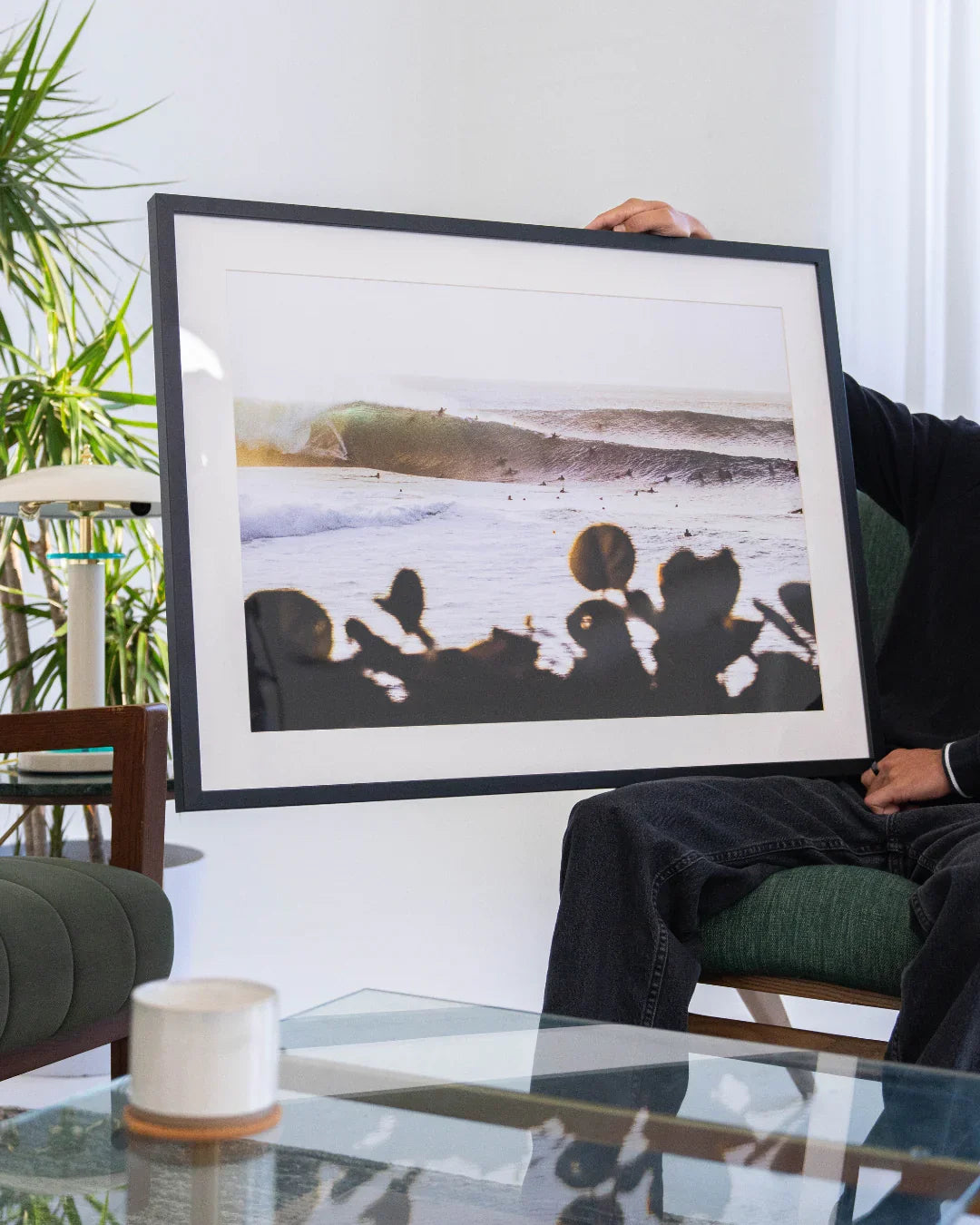 Person holding framed surf photography featuring ocean waves and surfers indoors with green armchair and plants