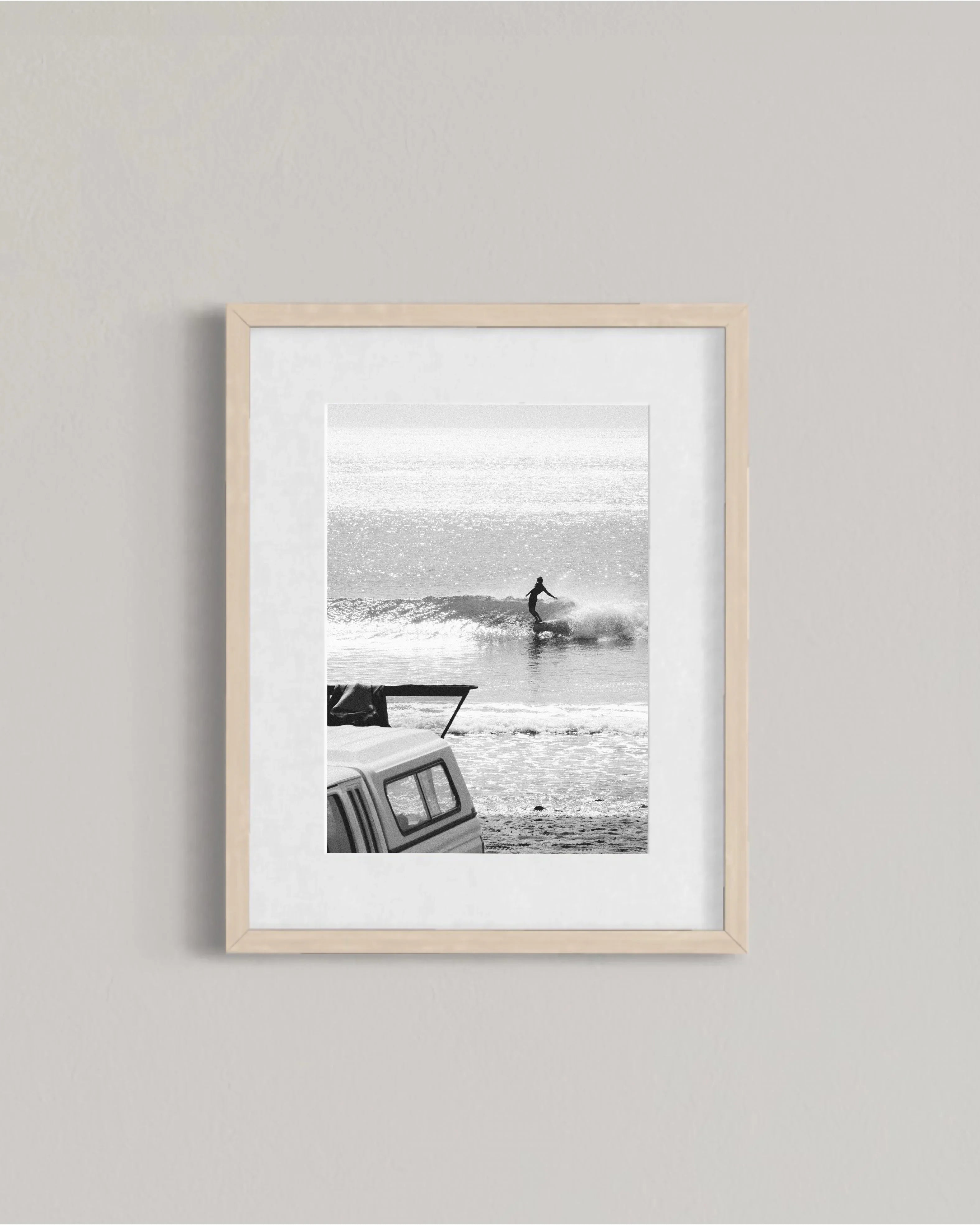 Black and white framed photo of surfer riding a wave with a parked camper on sandy beach
