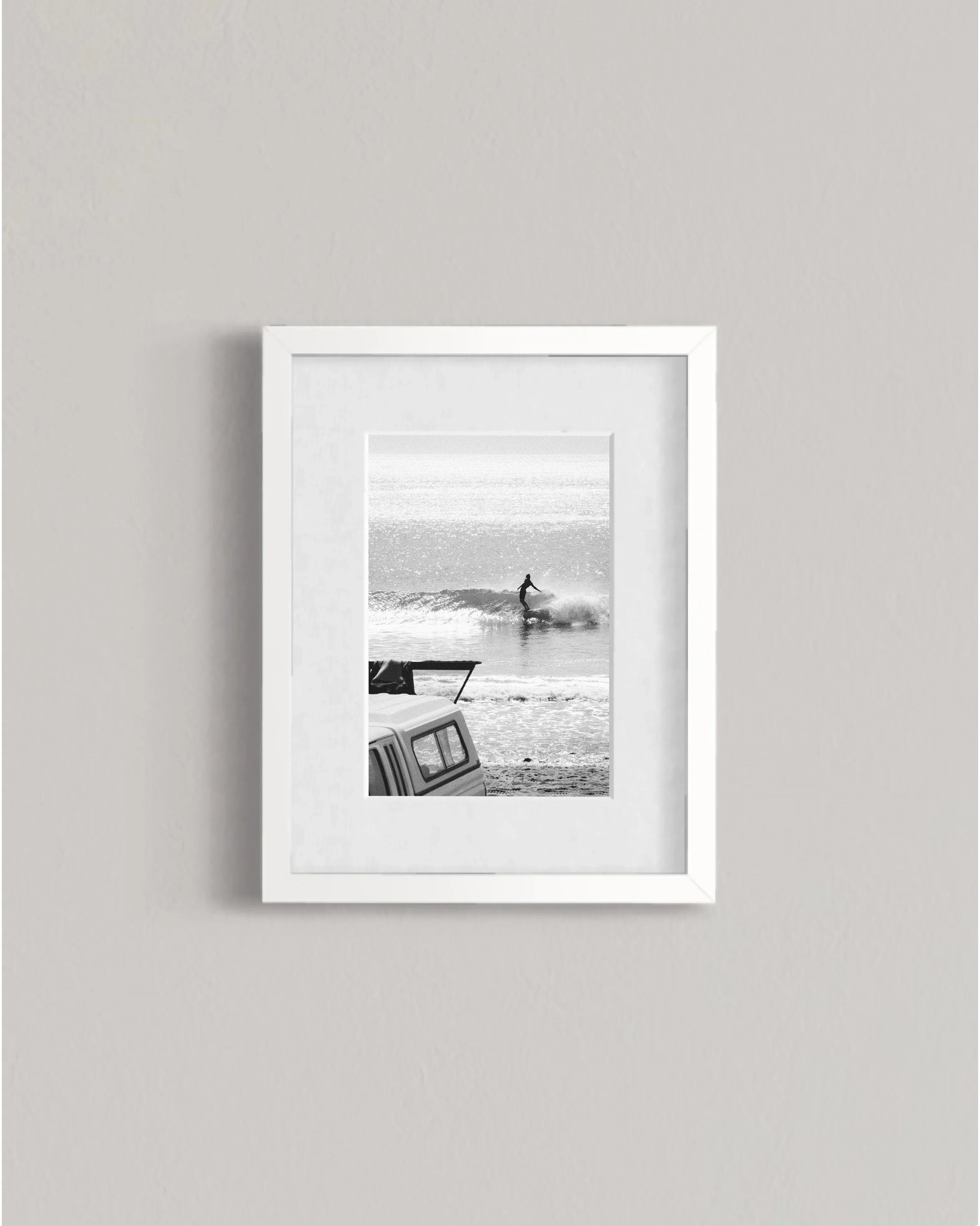 Black and white photo of a surfer riding a wave with a parked car in the foreground, framed on a wall