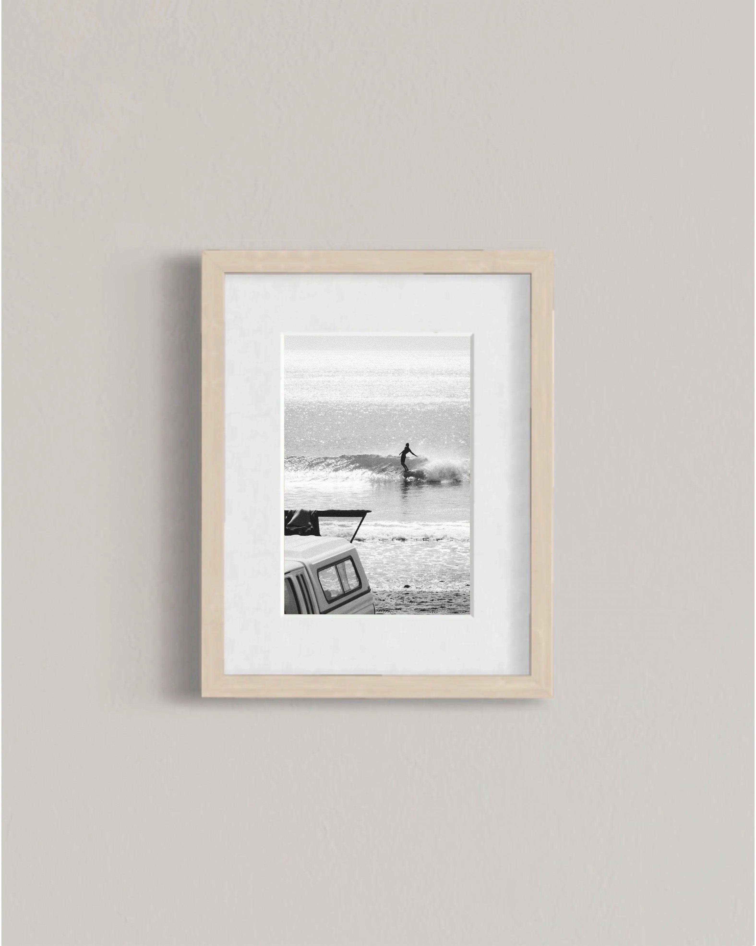 Black and white framed photo of surfer riding wave near beach with parked vehicle in foreground