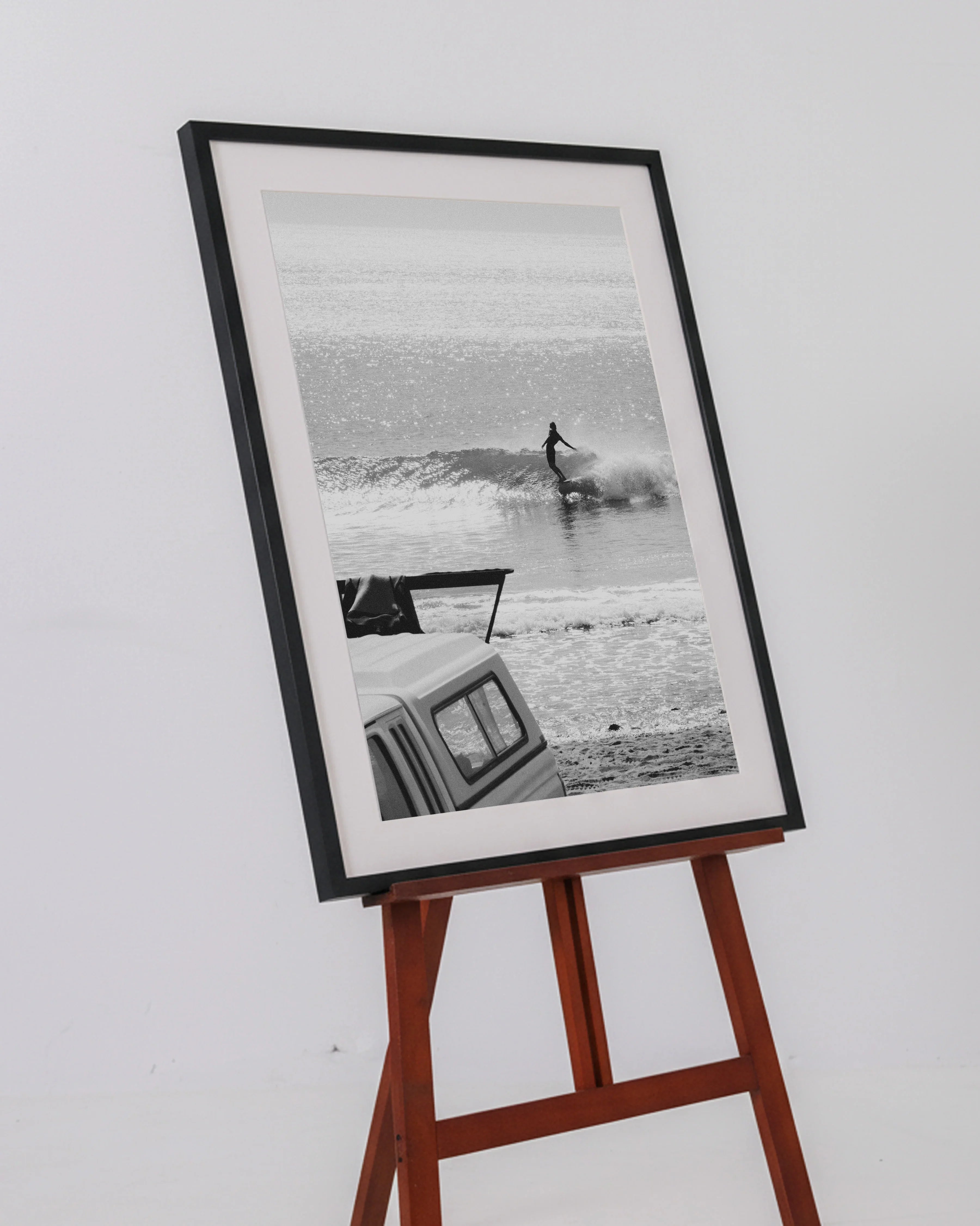 Black and white framed photo on wooden easel depicting surfer riding wave near beach and parked vehicle