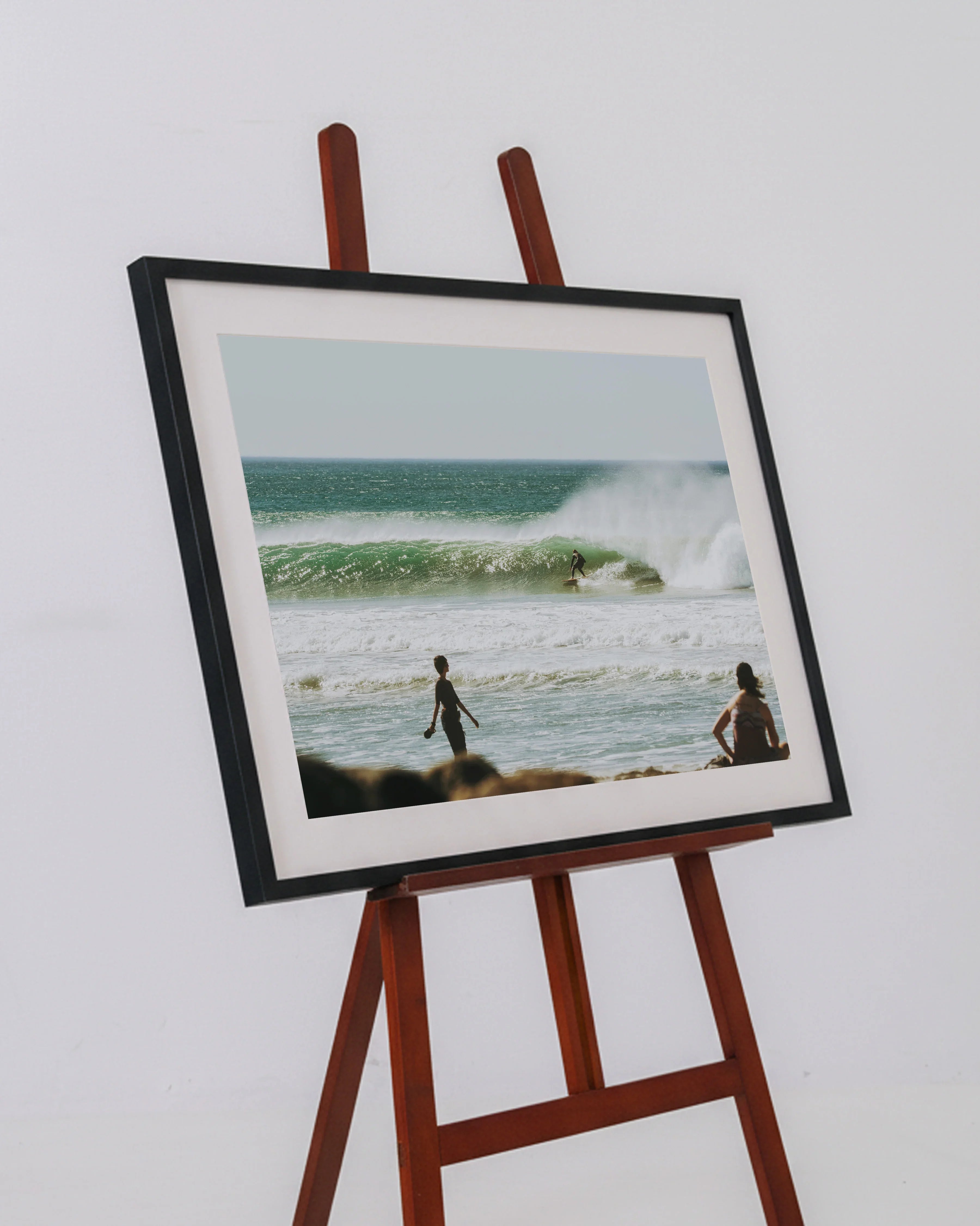 Framed photo on wooden easel showing surfer riding wave by beach with people watching