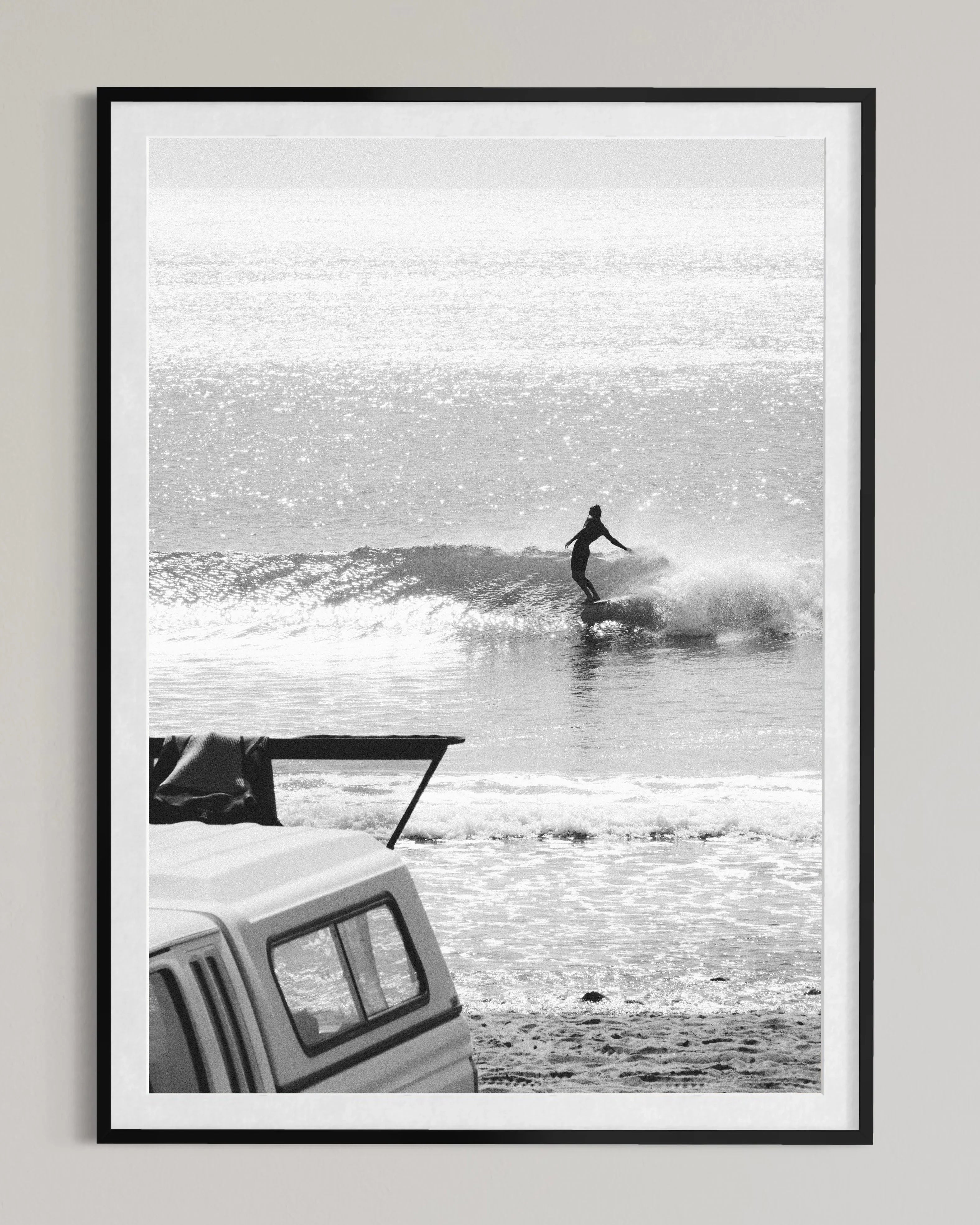 Black and white photo of a surfer riding a wave near a beach vehicle with surfboard rack