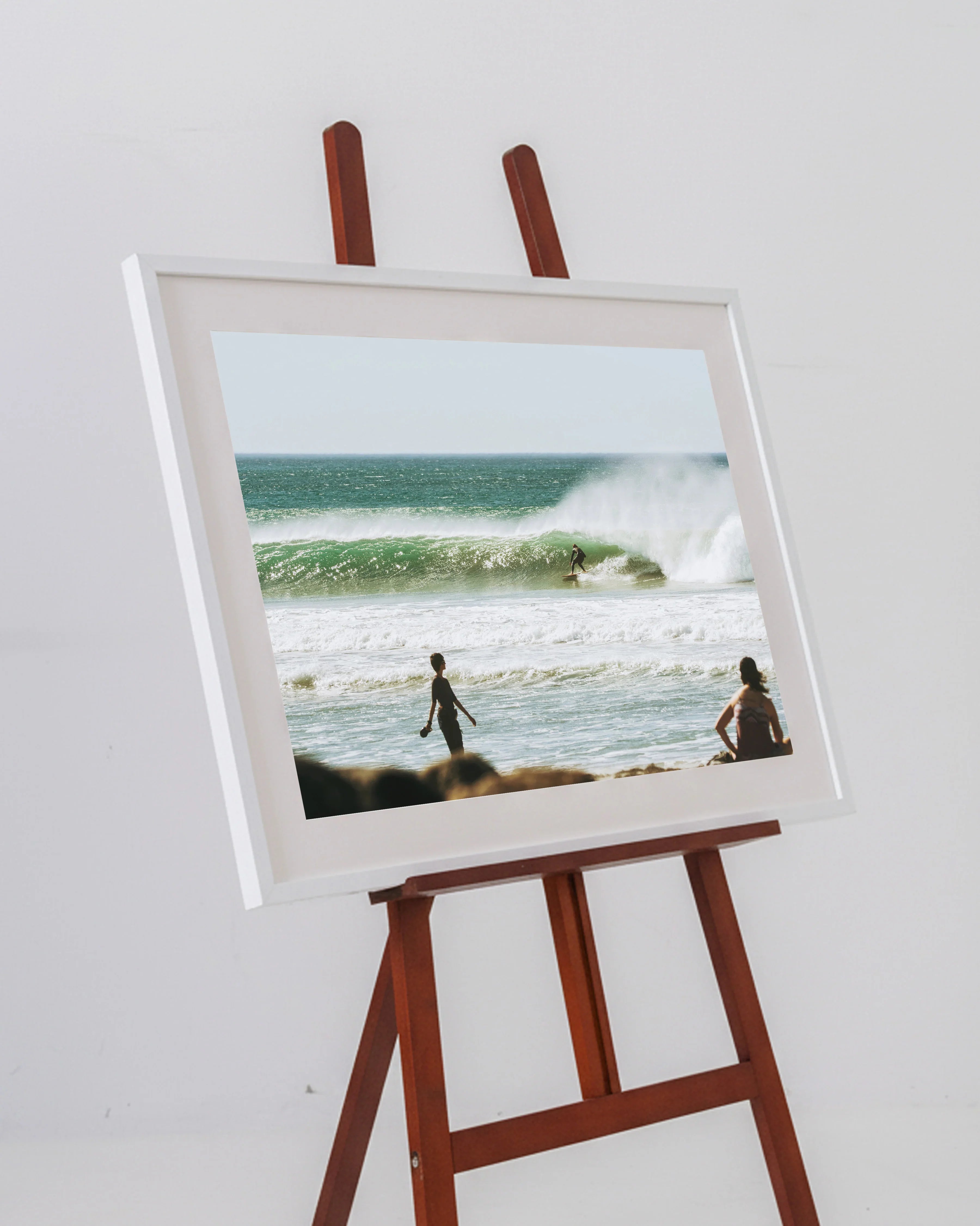 Framed photo on wooden easel showing surfer riding ocean wave with two people watching from beach