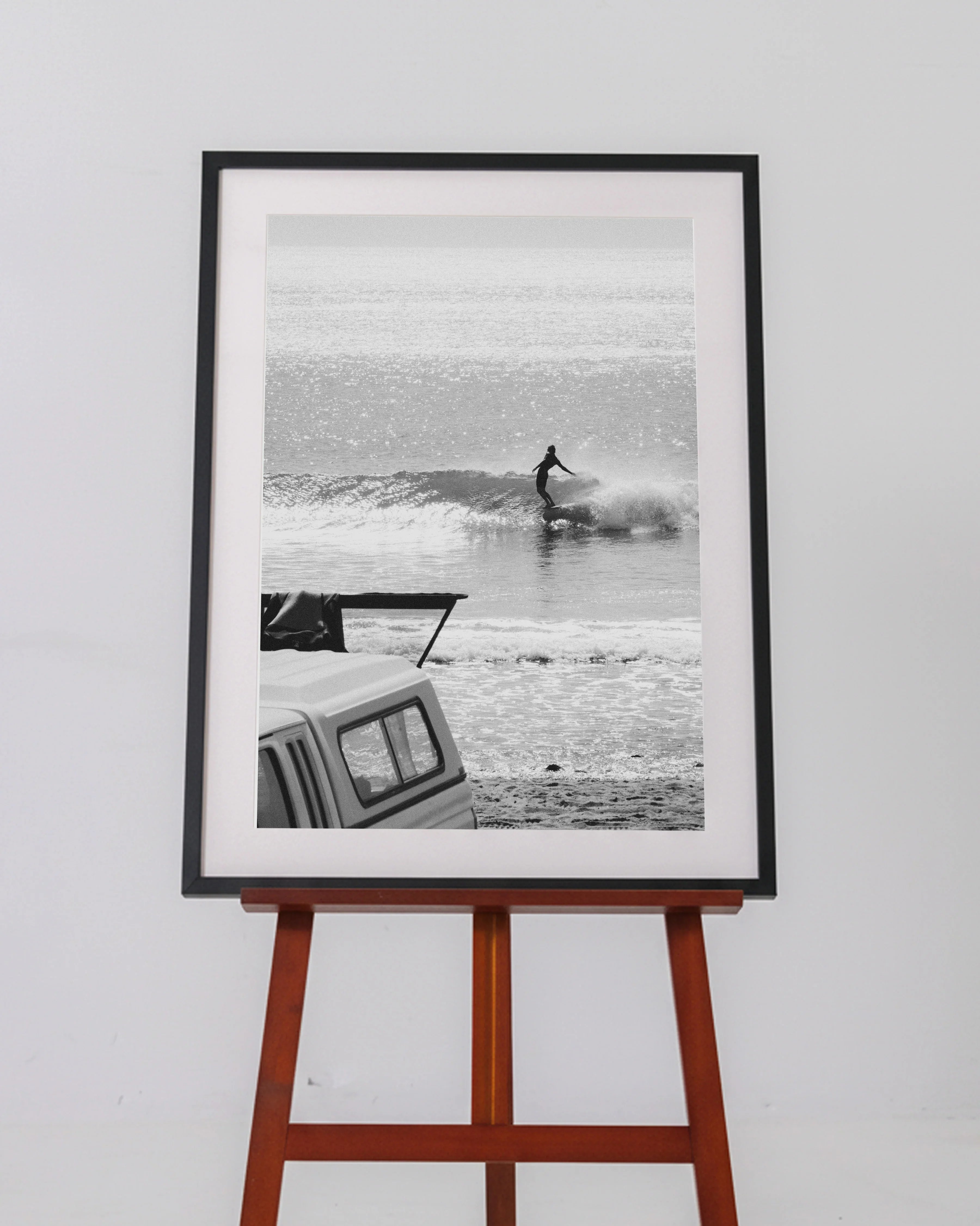 Black and white framed photo of a surfer riding a wave by the beach with a vehicle in foreground