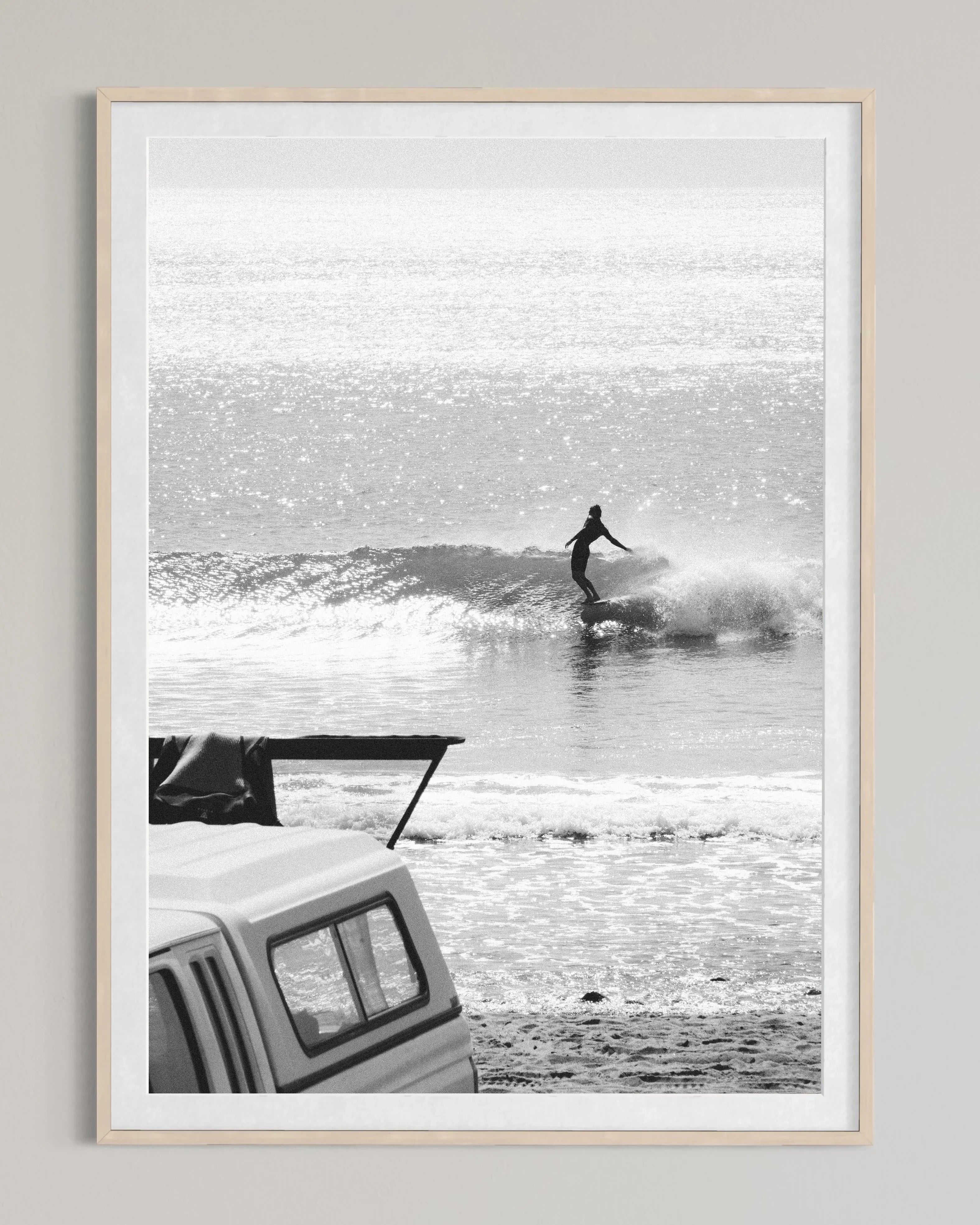 Black and white photo of a surfer riding a wave near a beach with a parked vehicle in the foreground