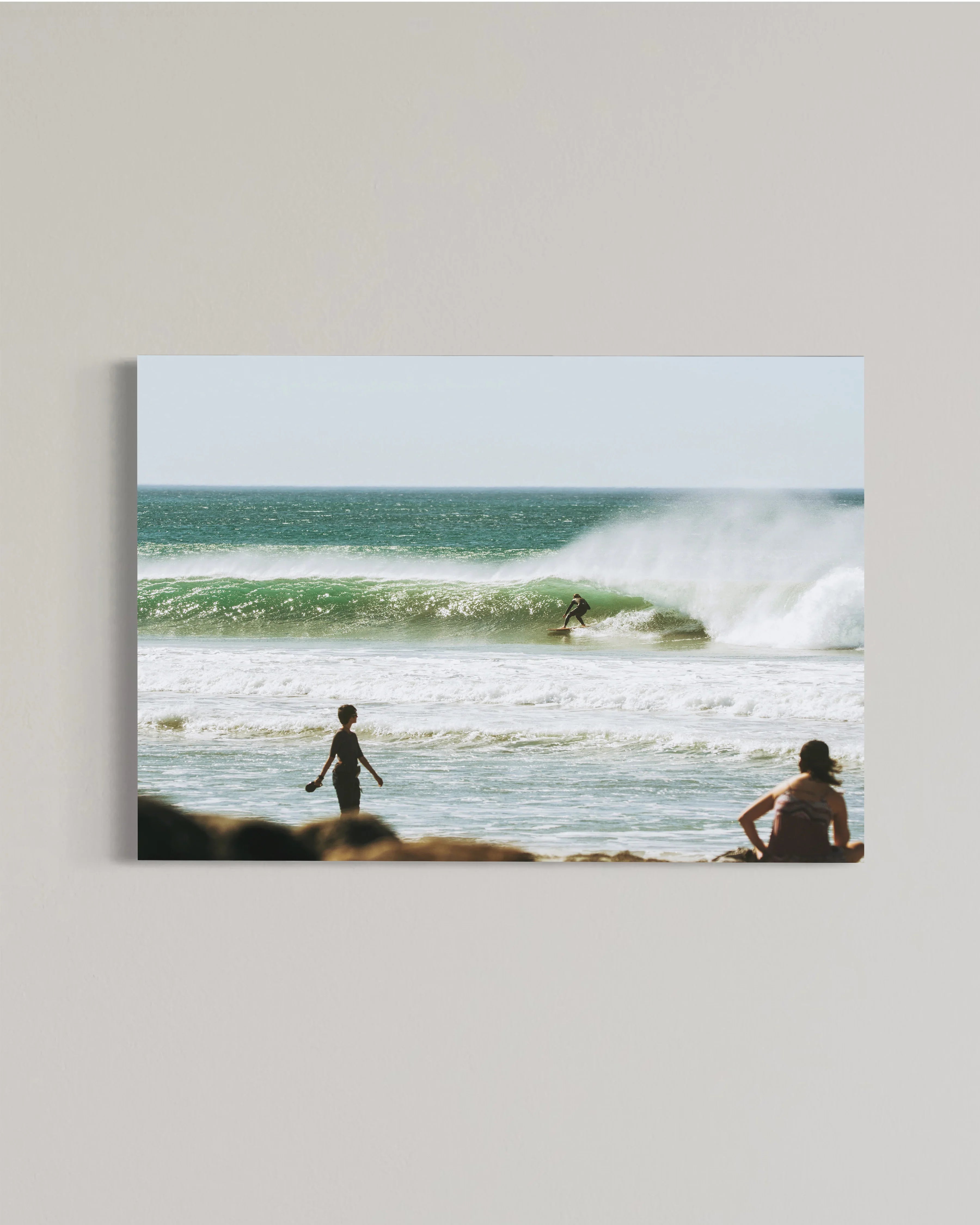 Surfer riding a green wave with beachgoers watching near the rocky shore on a sunny day