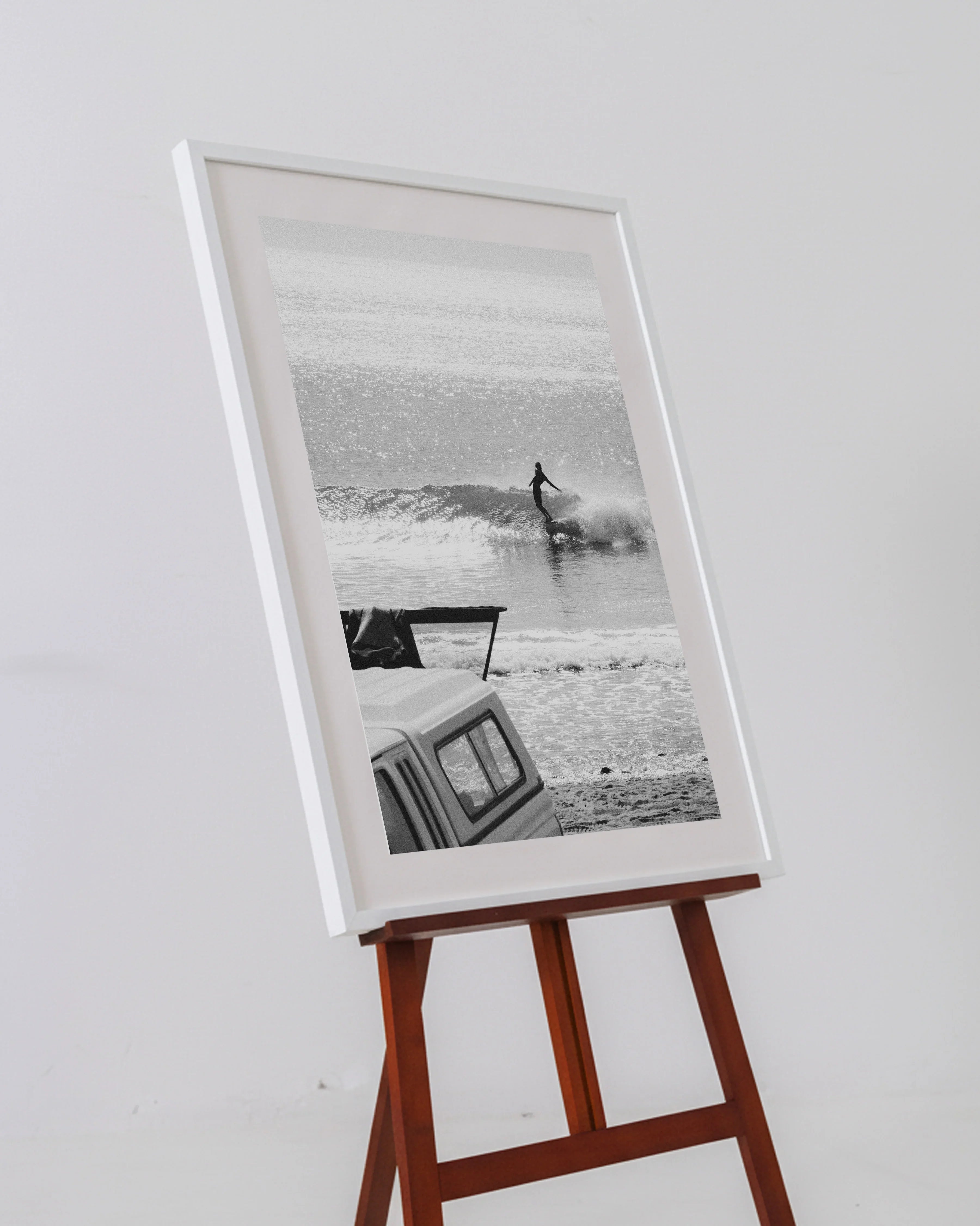 Black and white framed photo of a surfer riding a wave near a beach vehicle on a wooden easel