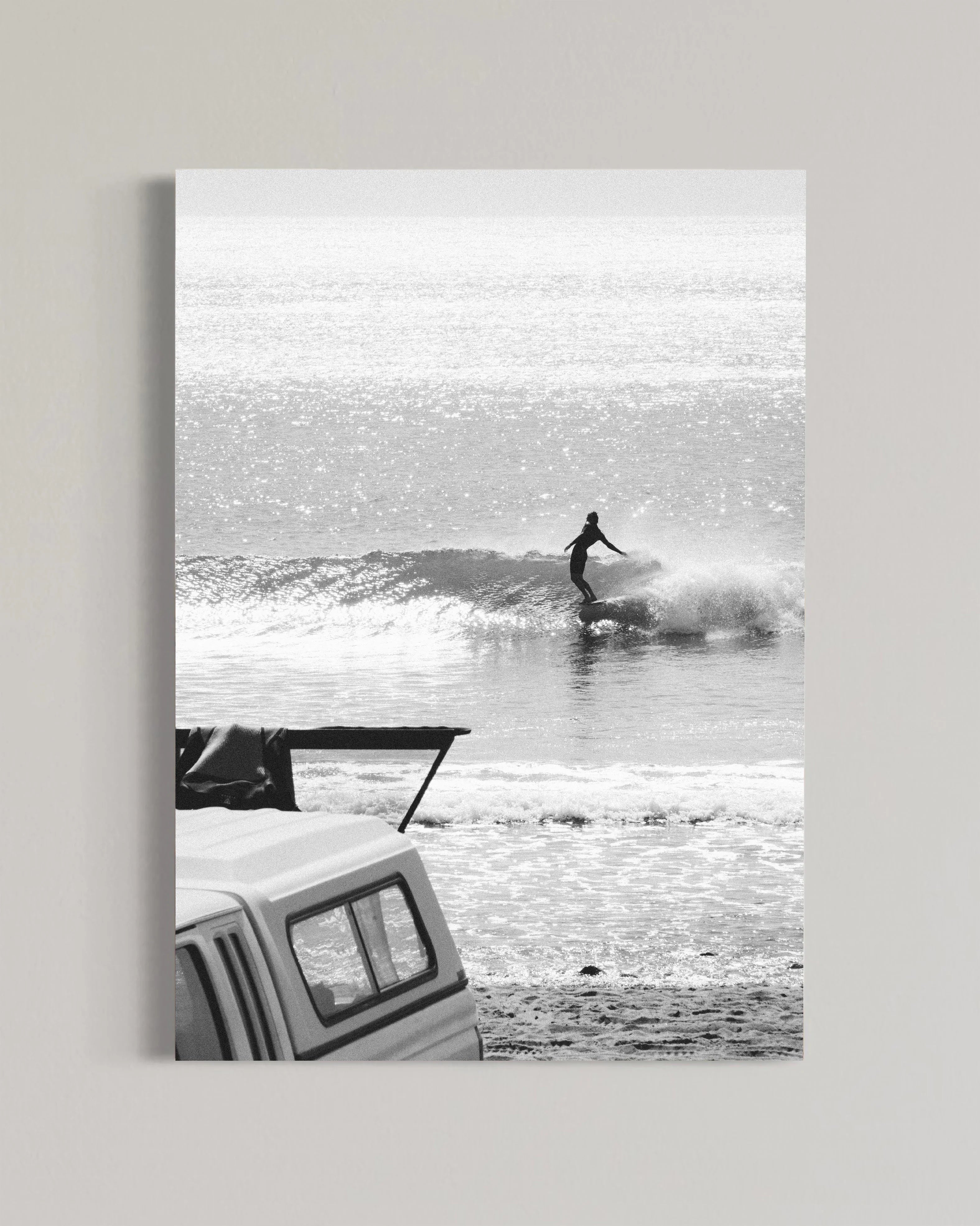 Black and white photo of a surfer riding a wave with a white pickup truck parked on the beach