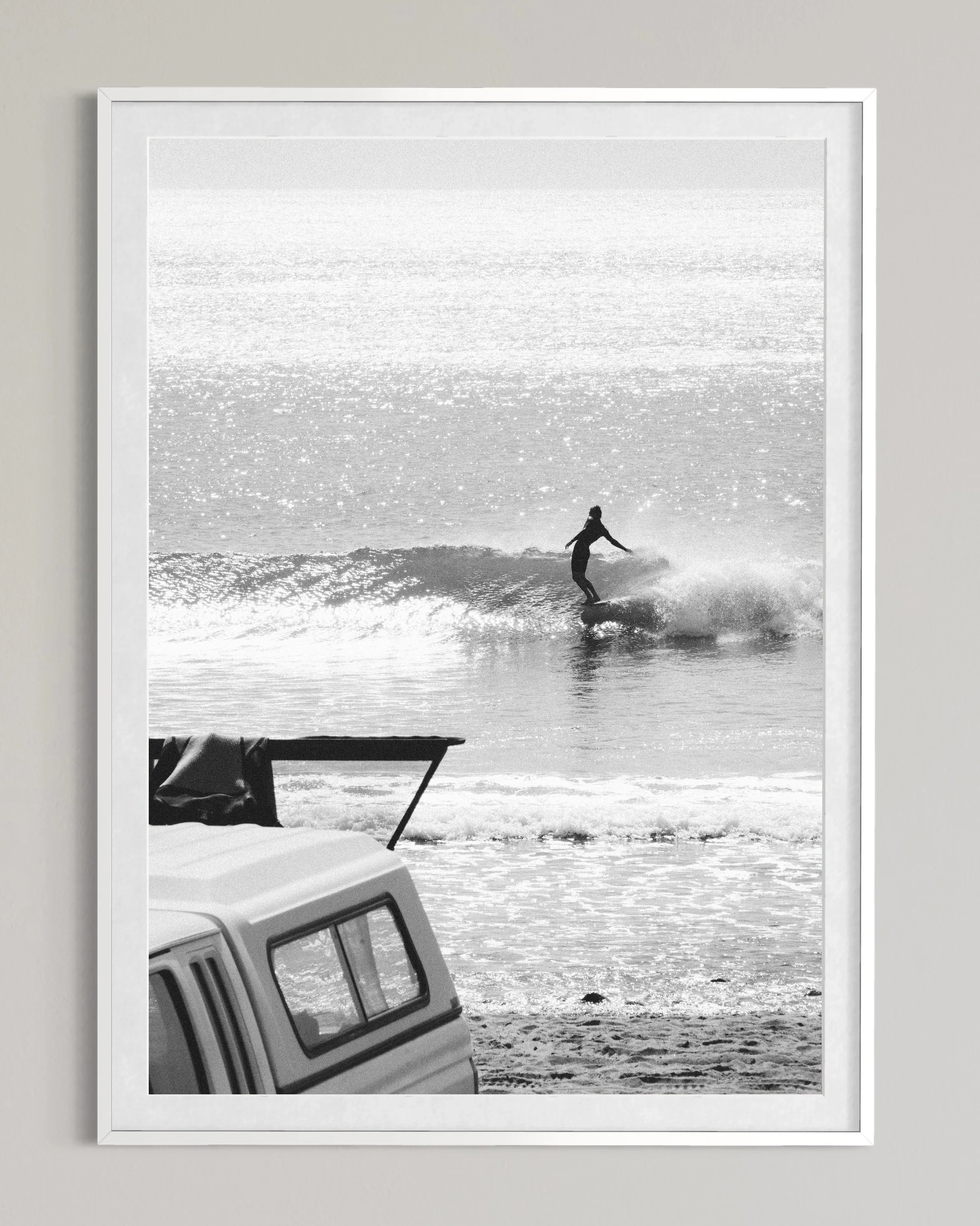 Black and white photo of a surfer riding a wave near a sandy beach with a parked vehicle in foreground