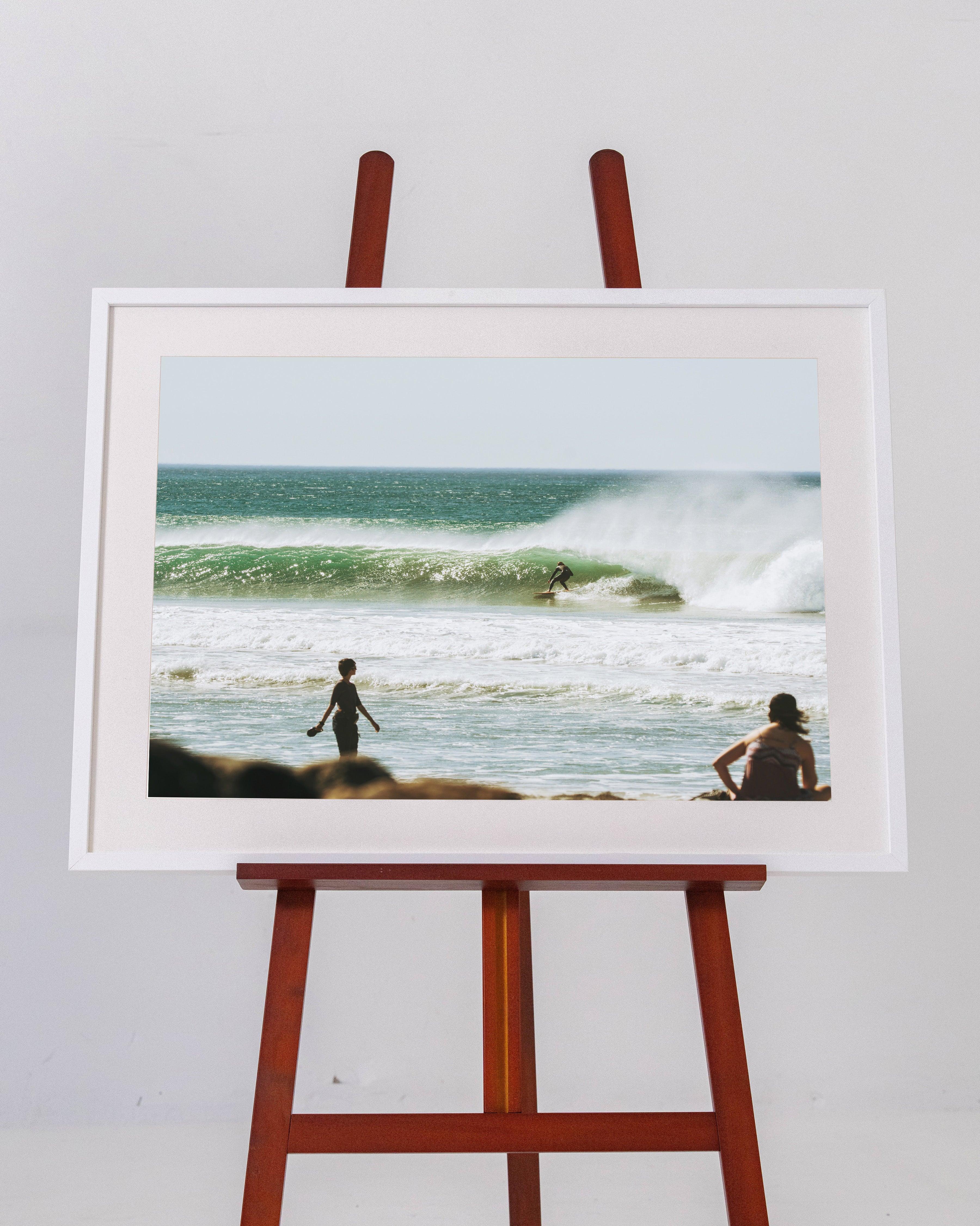Framed photo on wooden easel depicting surfer riding green wave with two people watching on beach