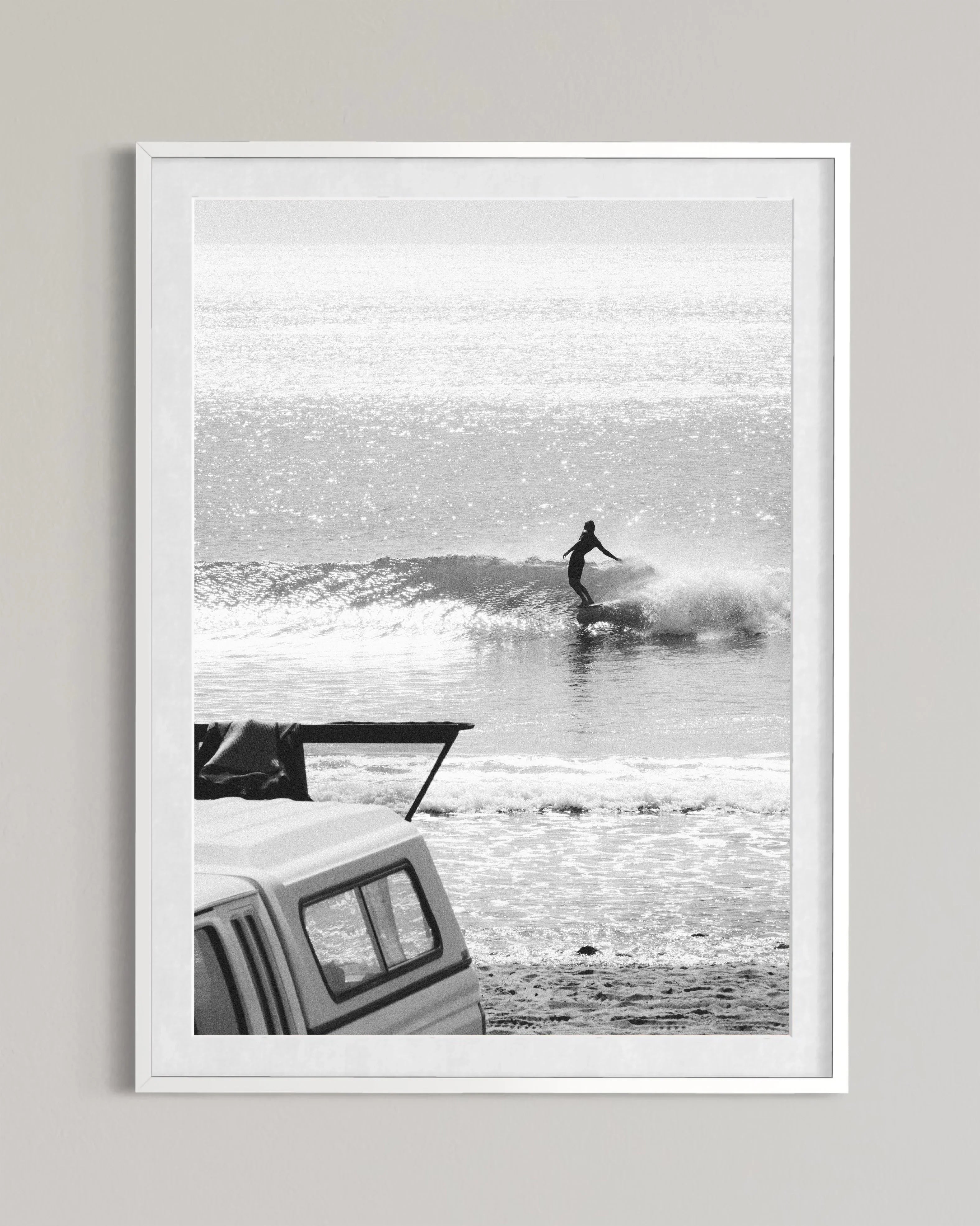 Black and white photo of surfer riding a wave with parked truck on sandy beach in foreground