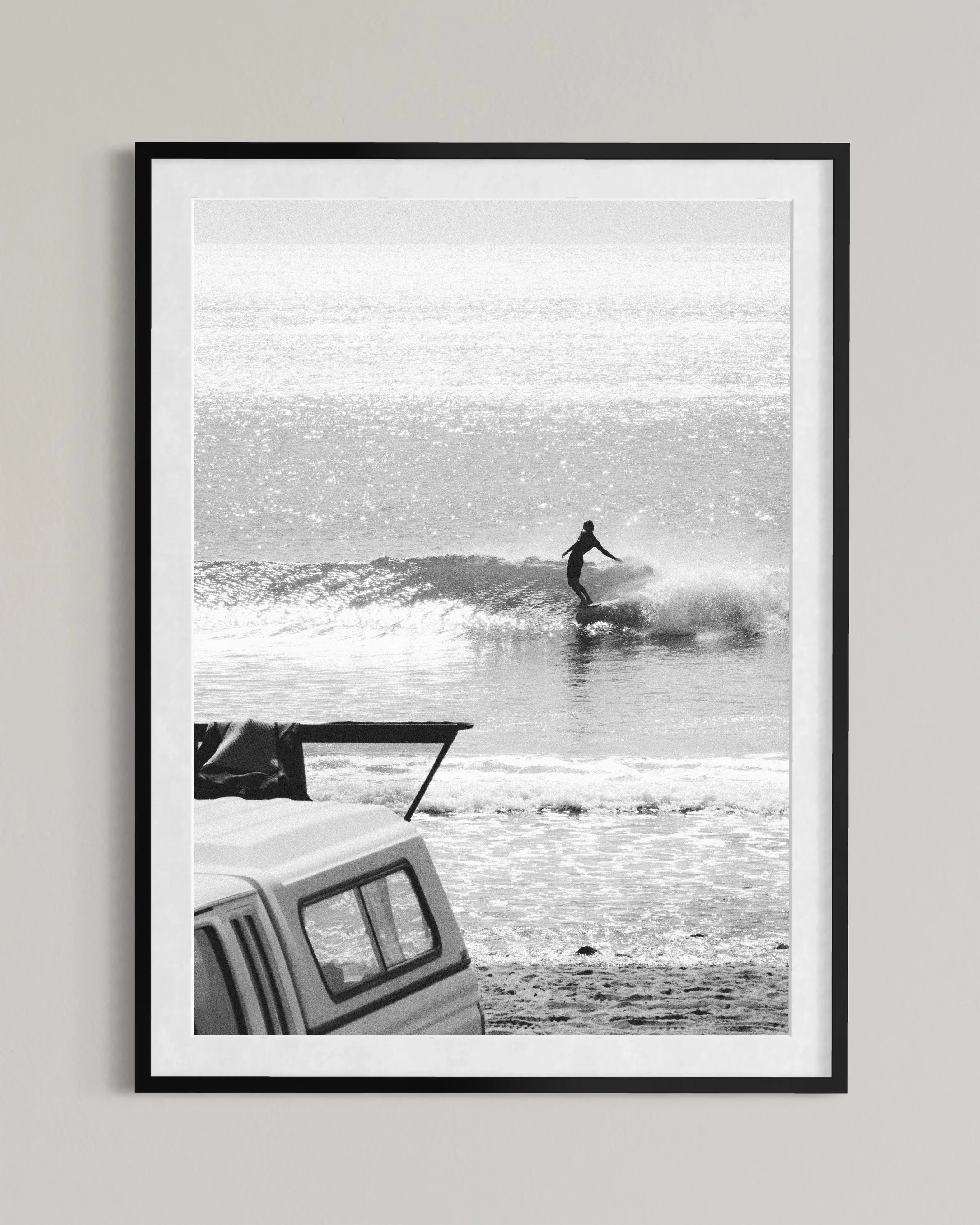 Black and white photo of surfer riding a wave with parked truck and surf rack on beach