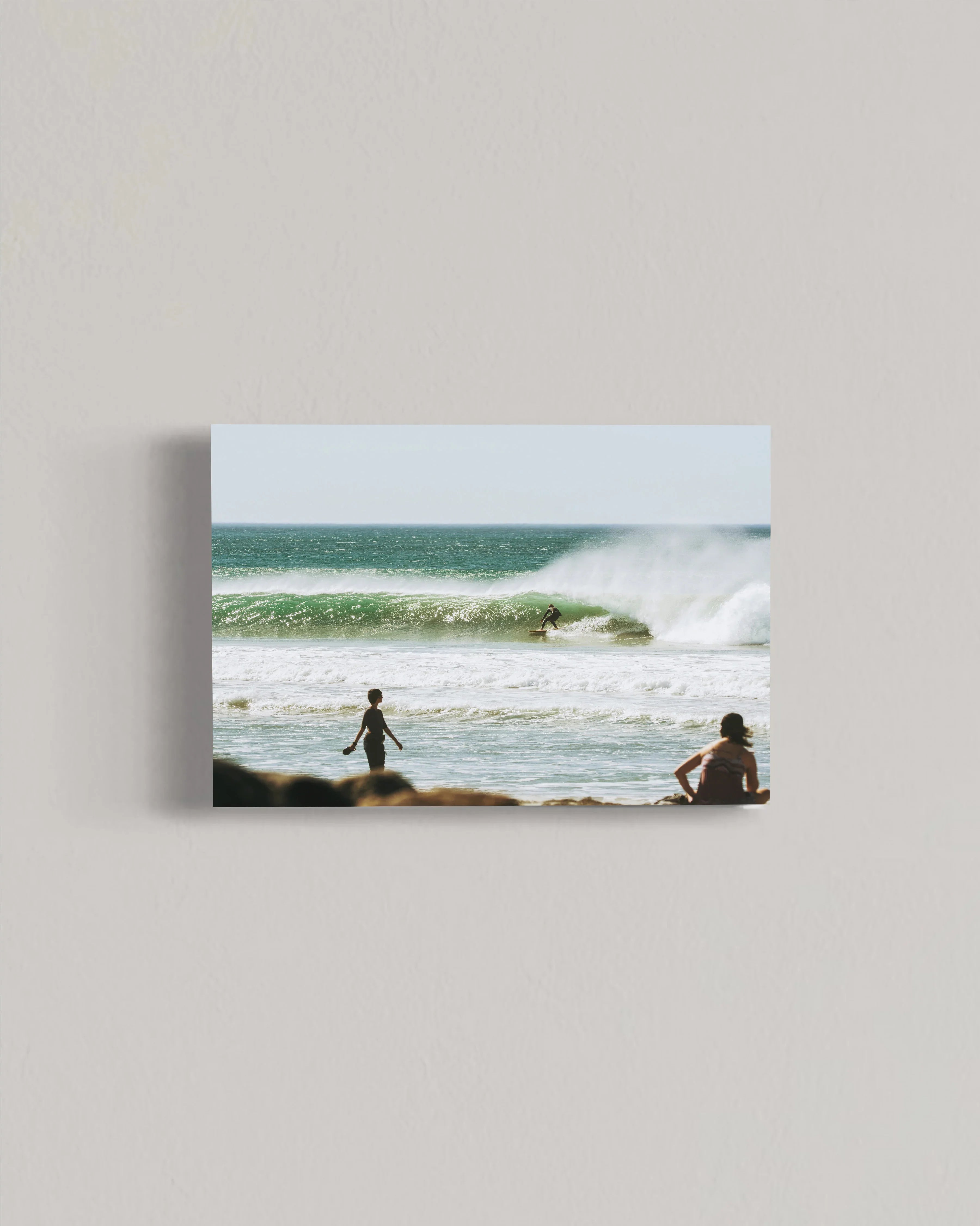 Surfer riding a green wave near beach with two people watching from shore on a sunny day