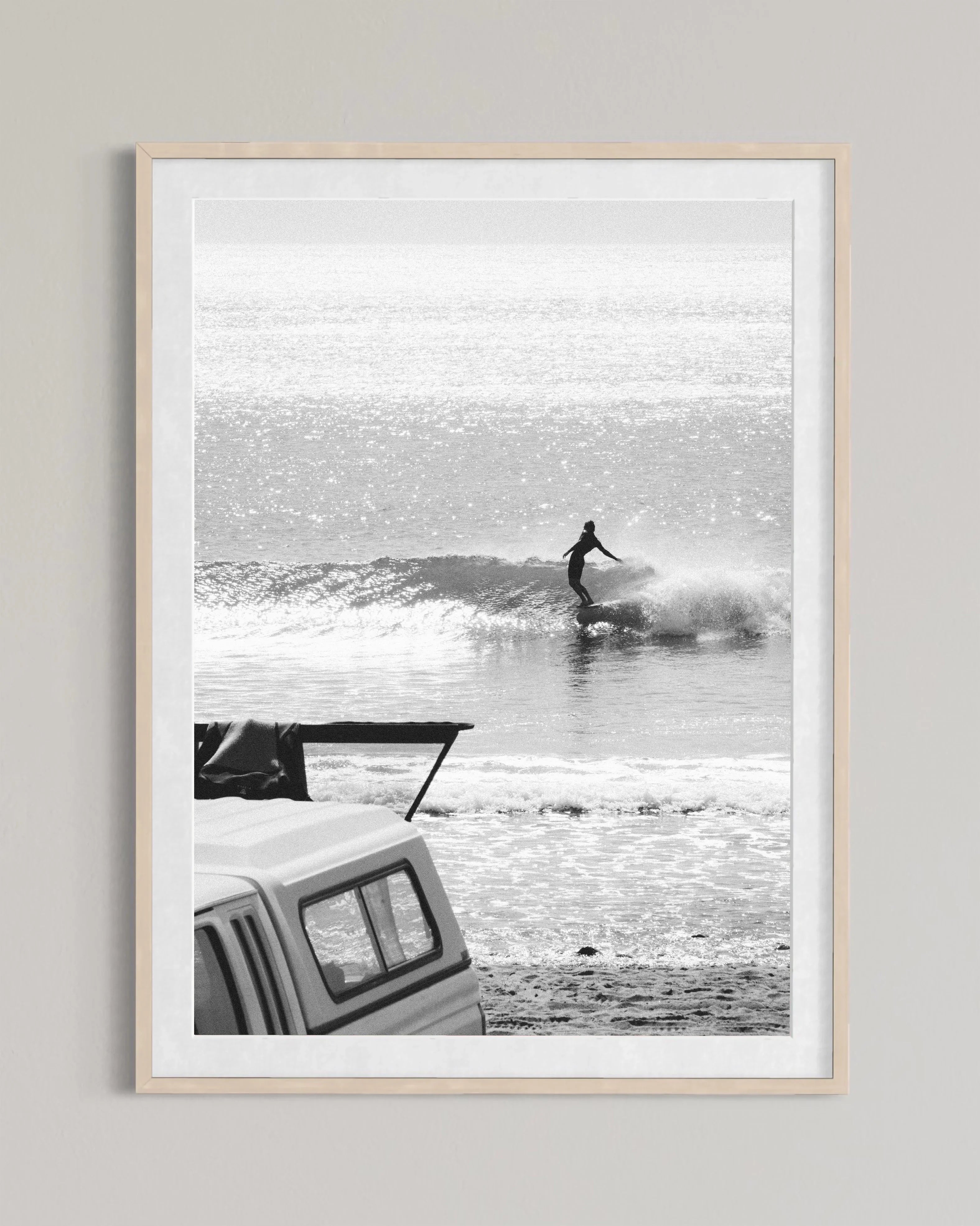 Black and white photo of a surfer riding a wave with a parked camper truck on the beach