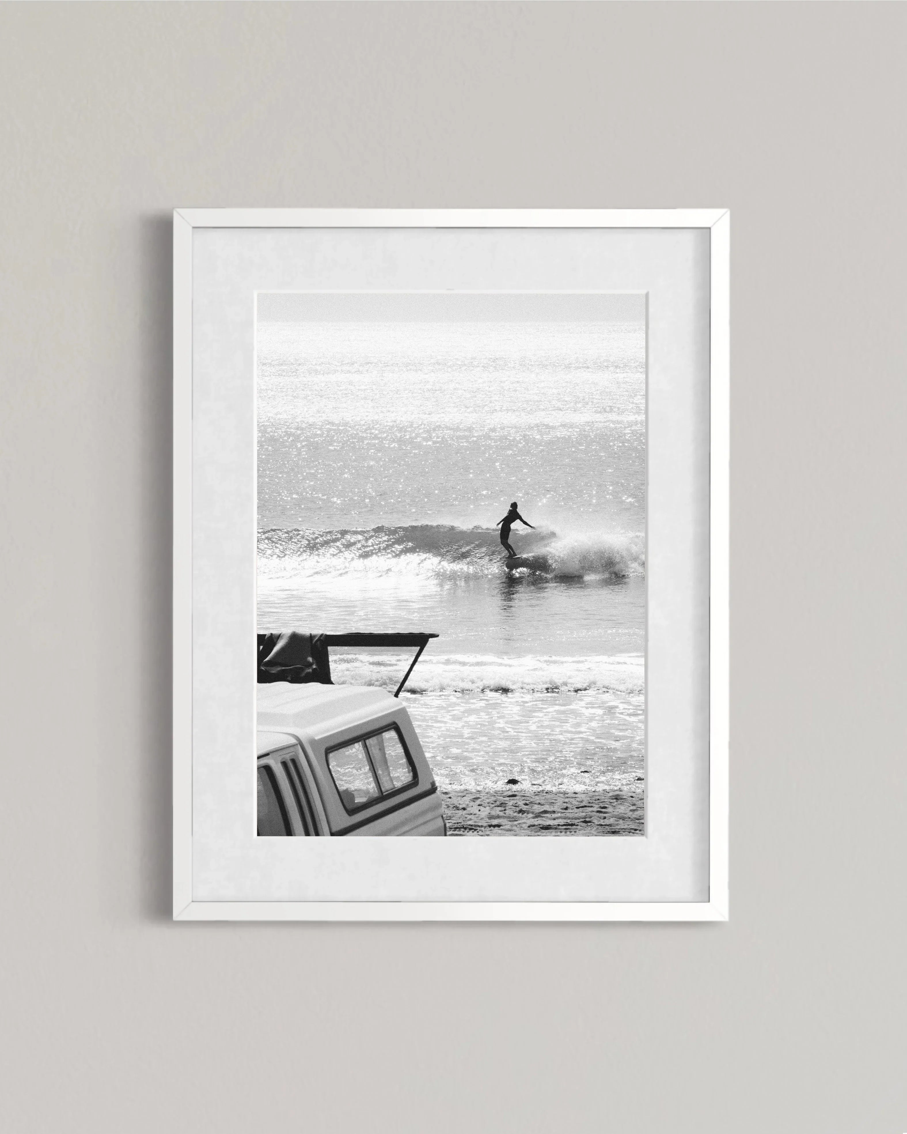 Black and white photo of a surfer riding a wave with a parked truck on the beach
