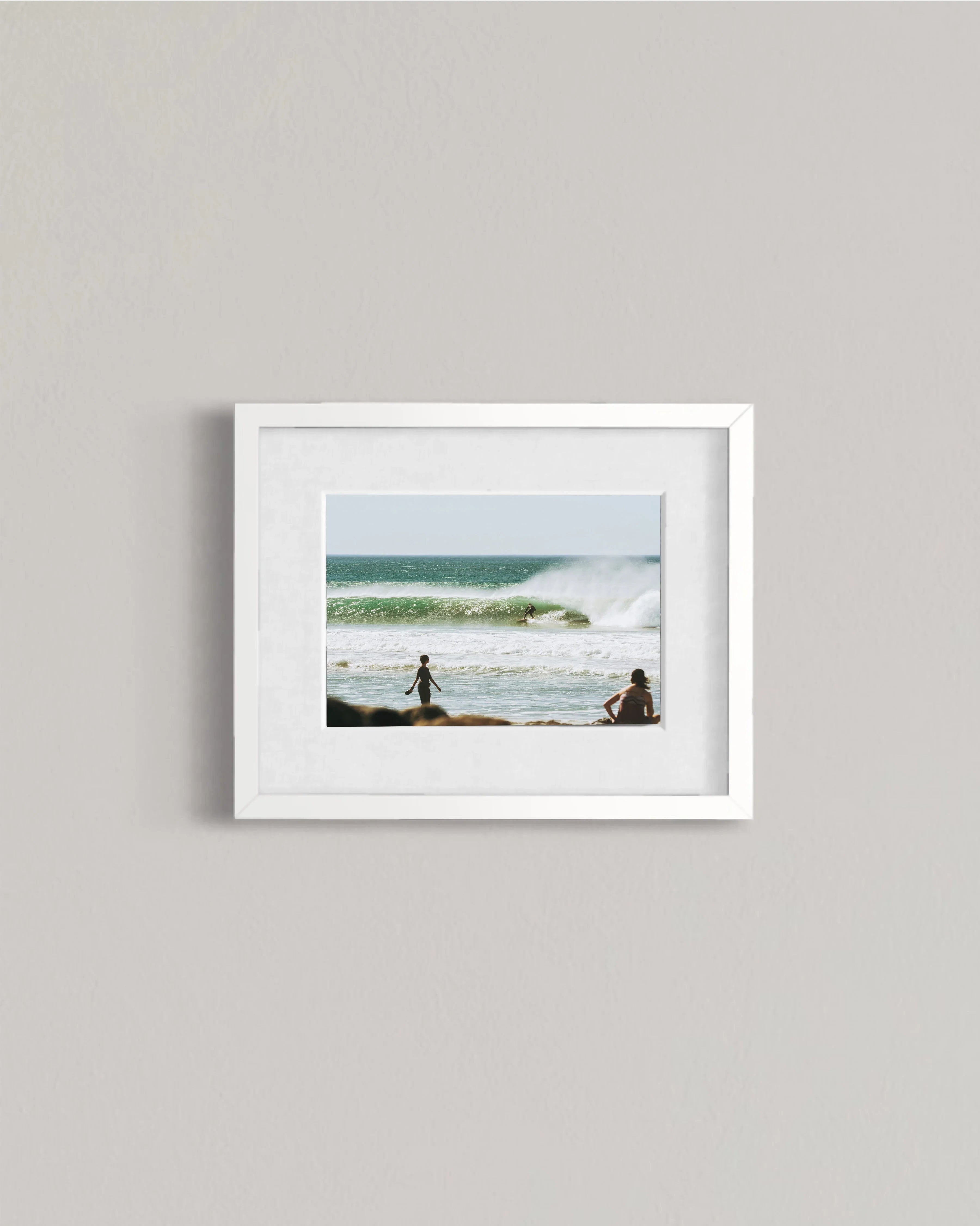 Framed photo of surfers and beachgoers by ocean waves under blue sky on gray wall