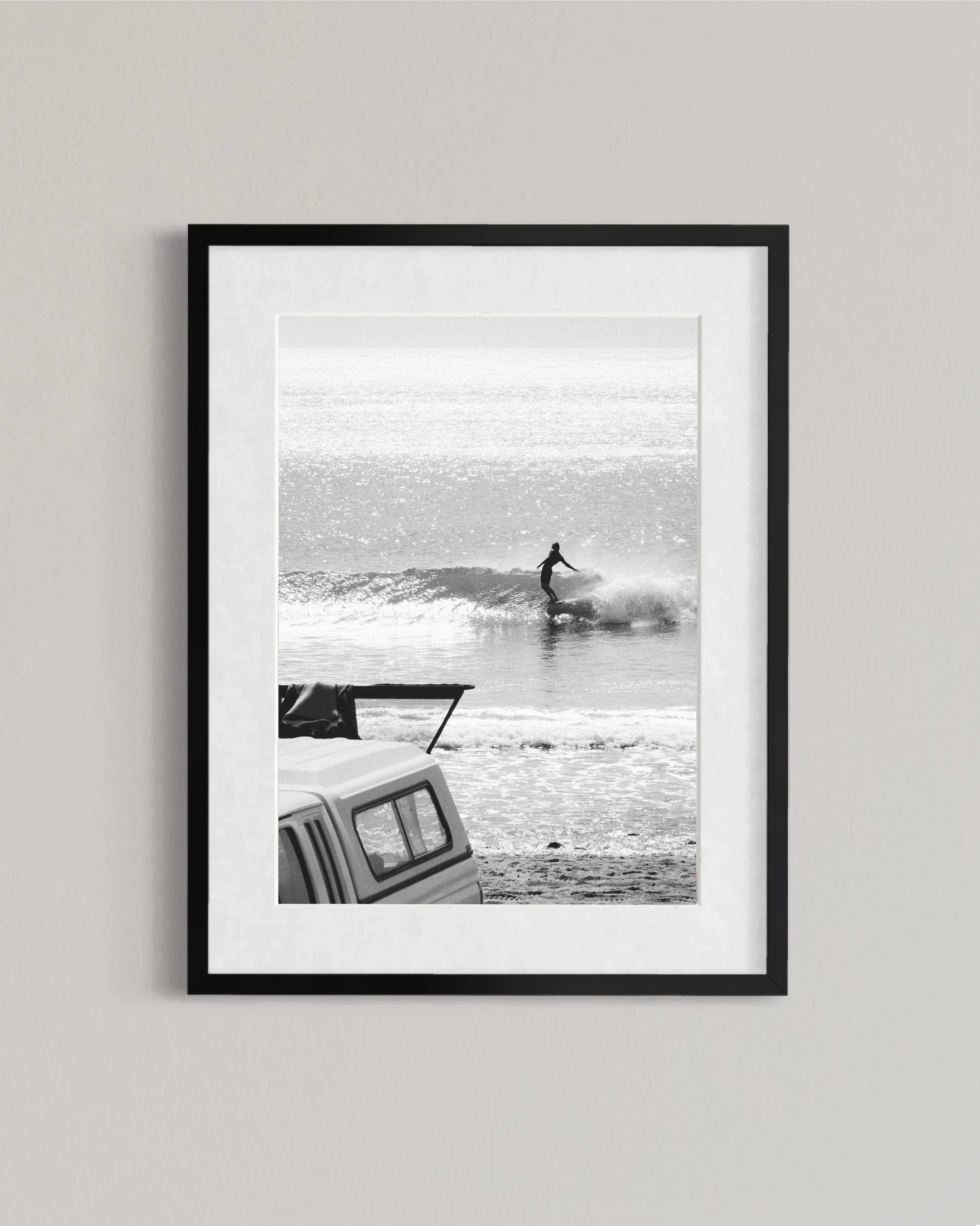 Black and white framed photo of a surfer riding a wave with a parked pickup truck on the beach
