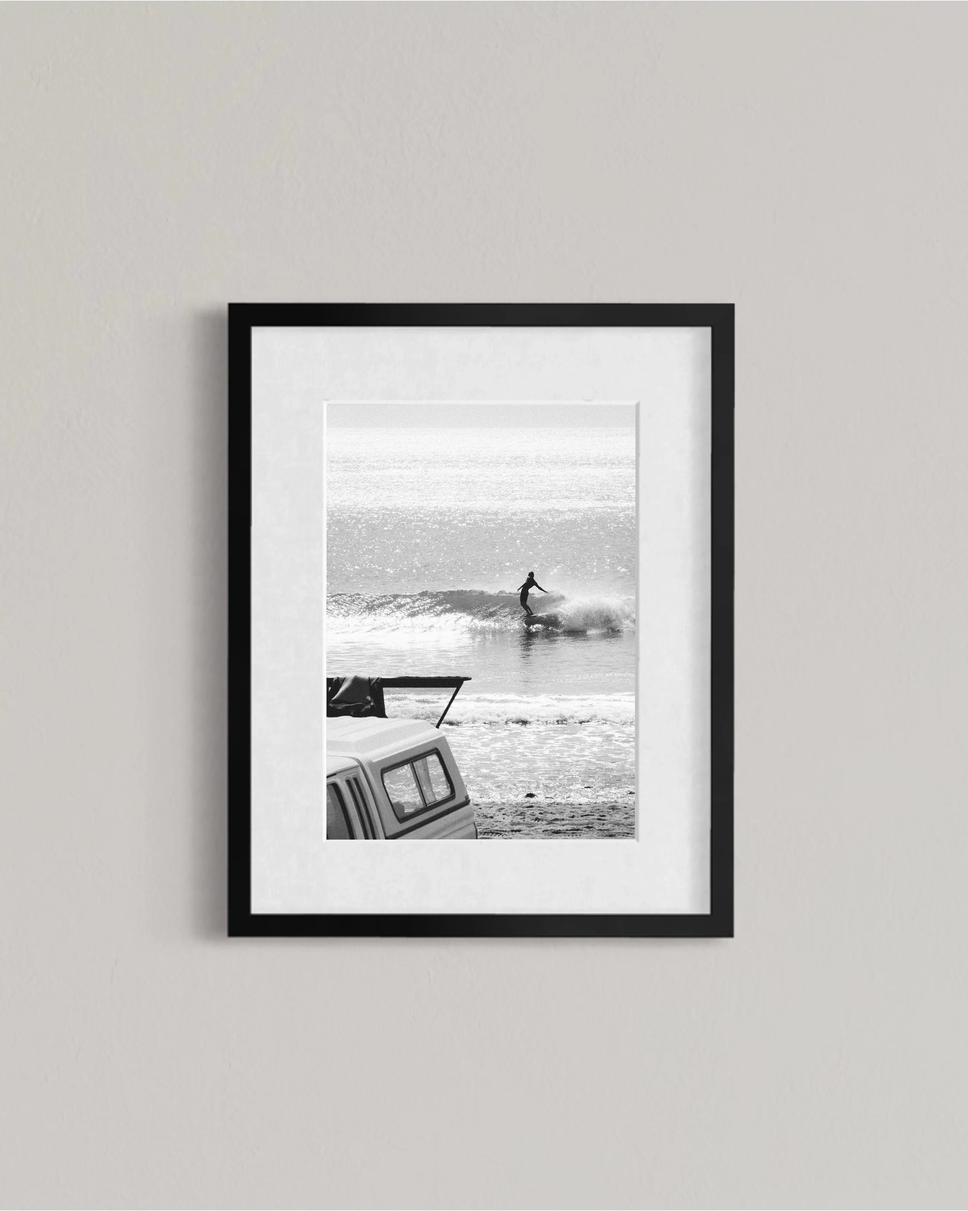 Black and white framed photo of a surfer riding a wave with a parked vehicle on a beach