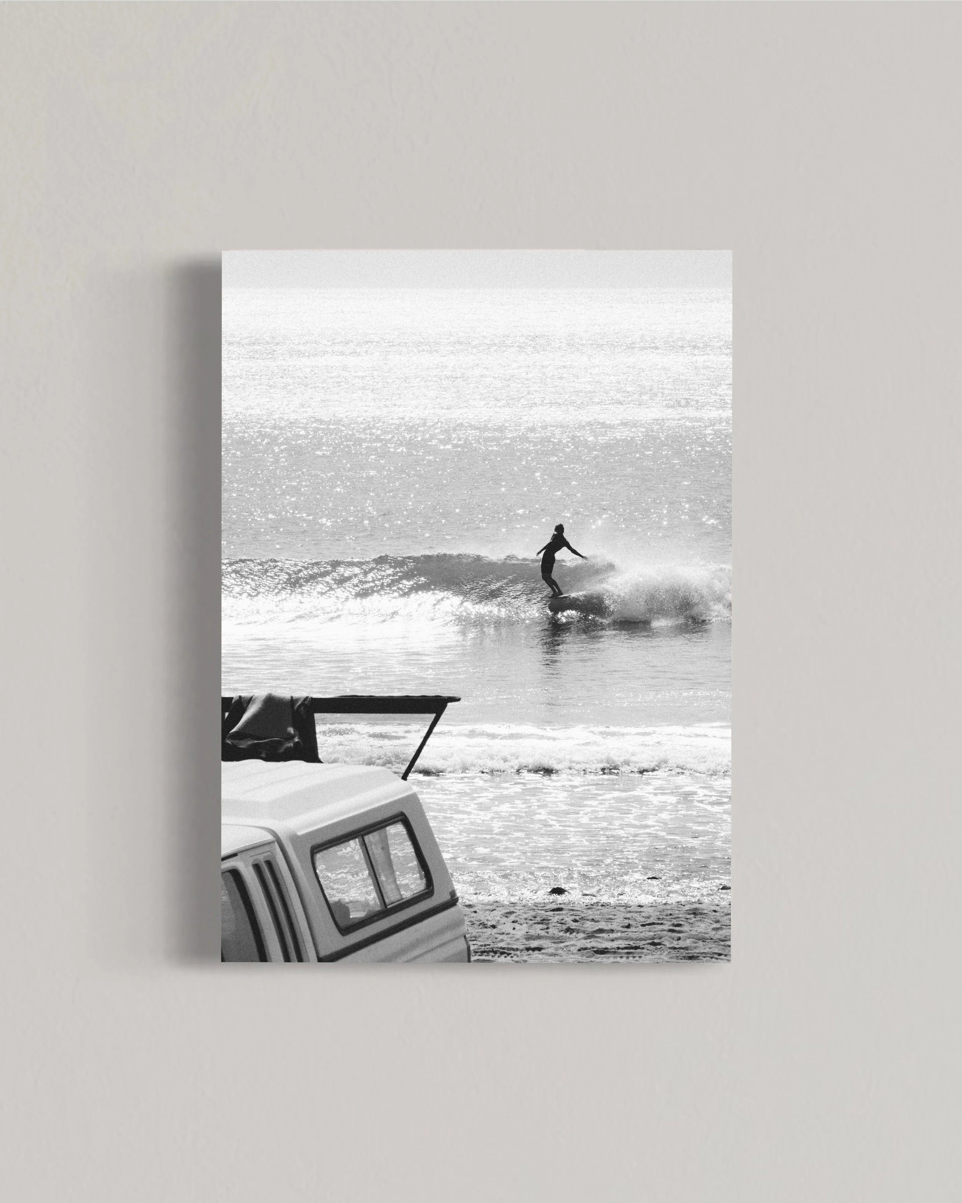 Black and white photo of surfer riding wave near beach with parked van and surfboard