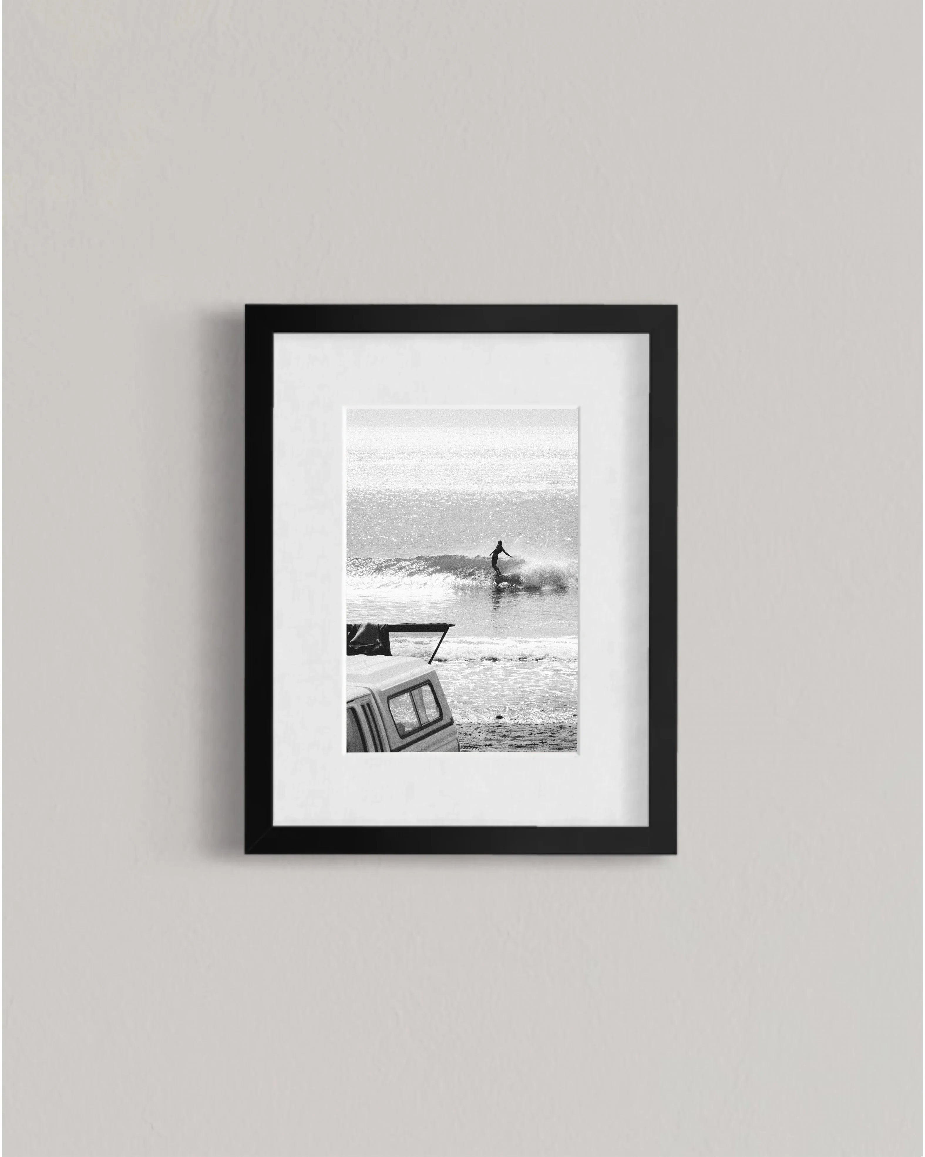 Black and white framed photo of surfer riding wave by beach with parked vehicle in foreground