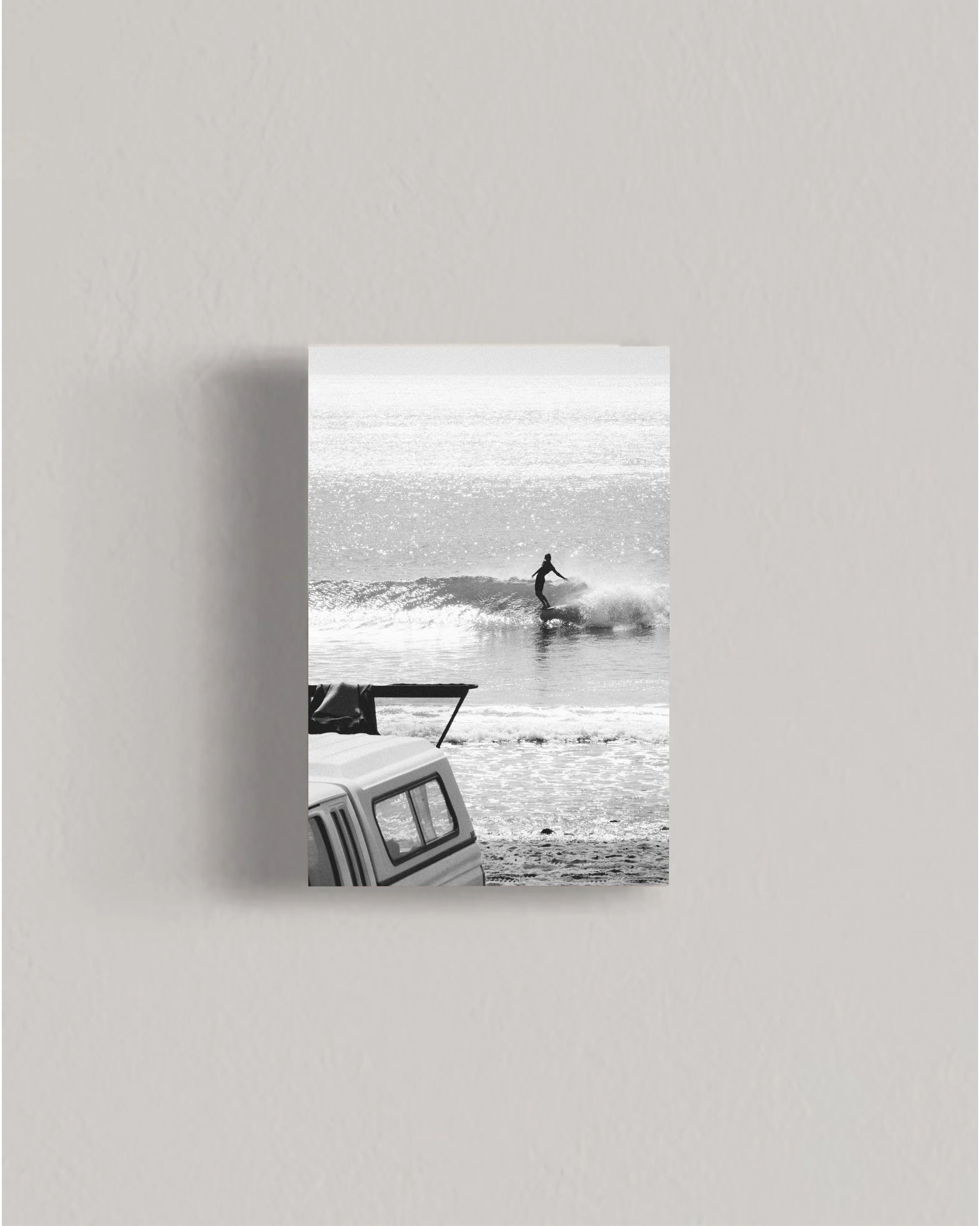 Black and white photo of a surfer riding a wave near a parked van on the beach