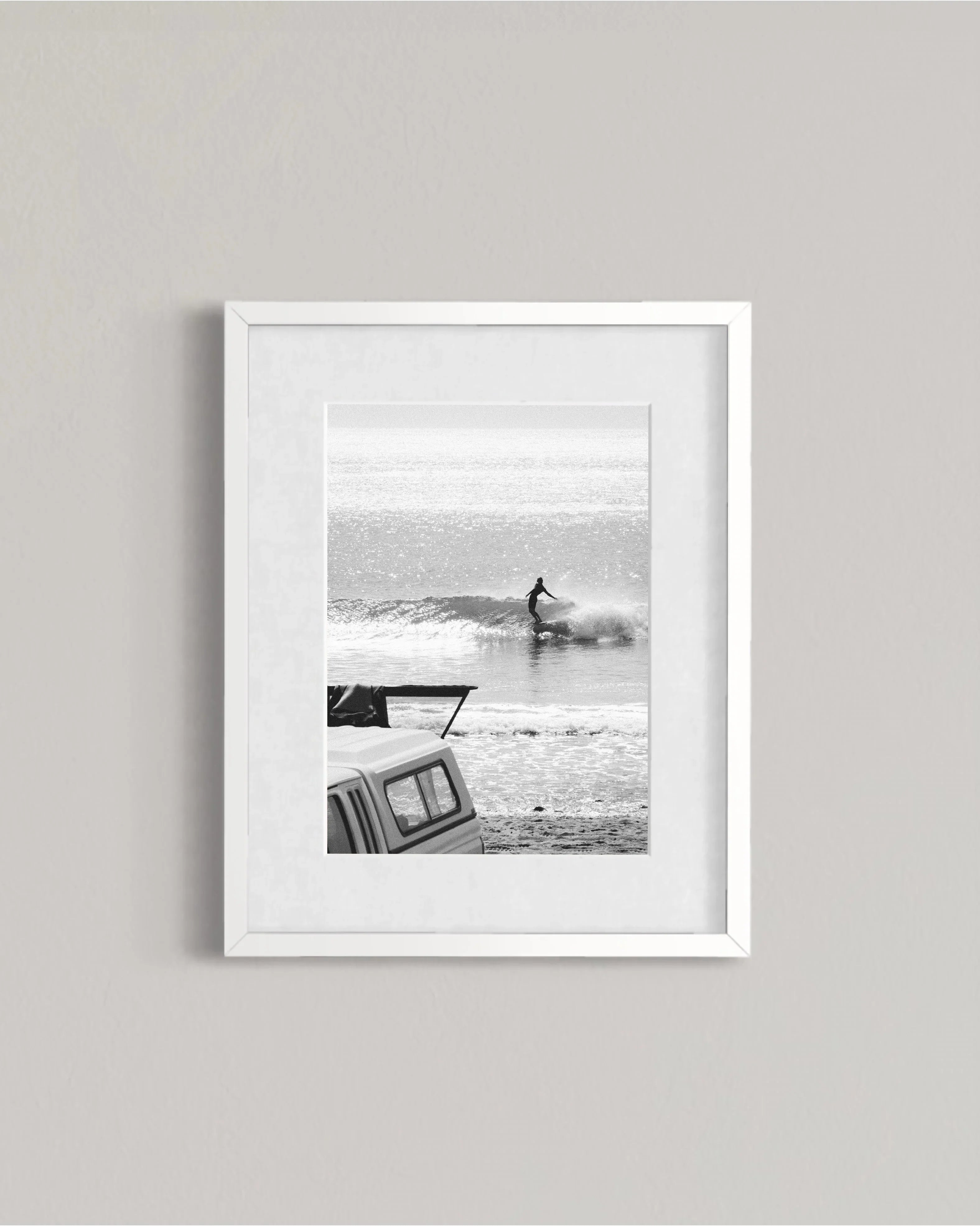 Black and white photo of a surfer riding a wave near a parked vehicle on the beach
